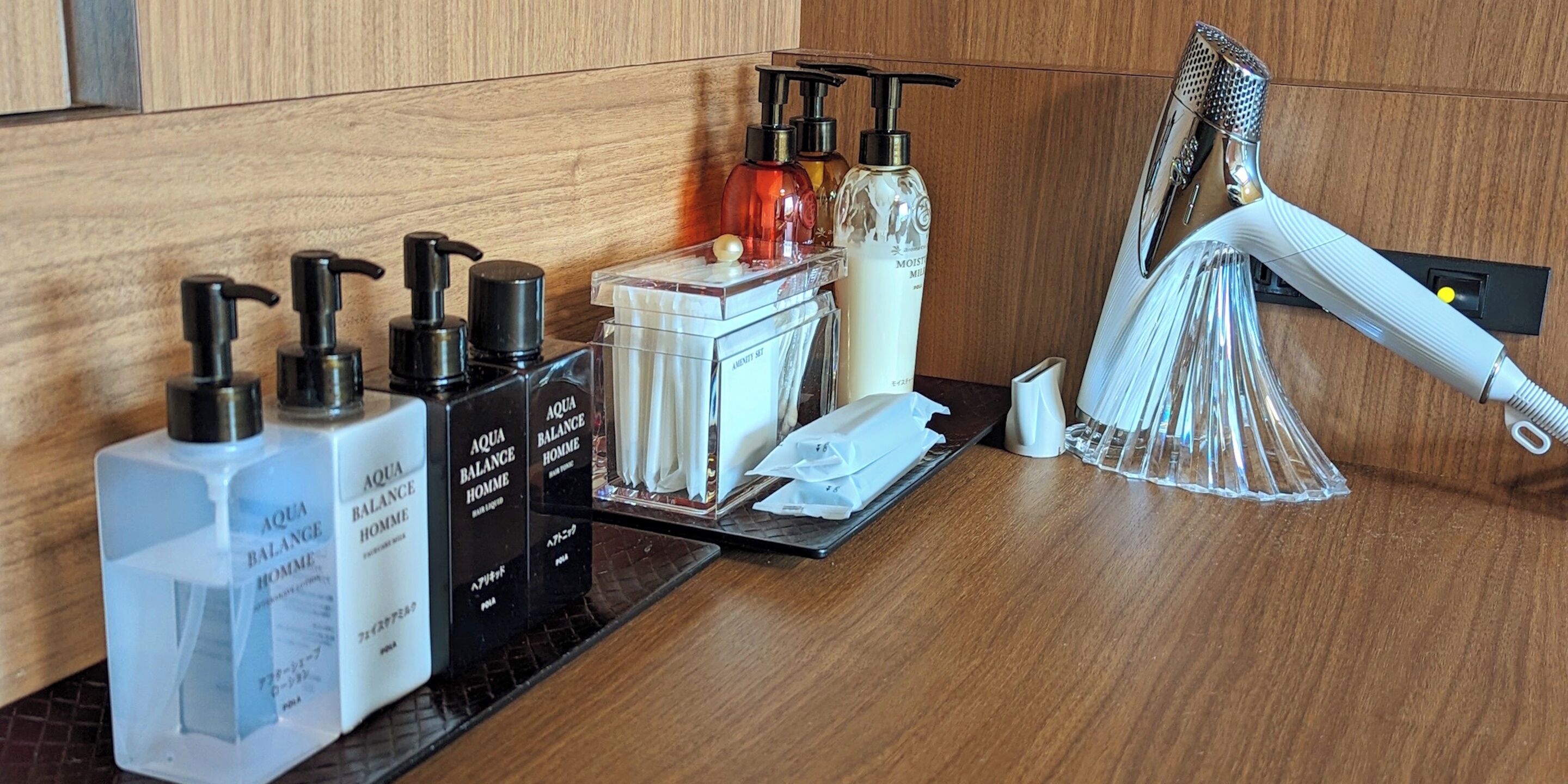 Un ripiano del bagno con articoli da toilette, tra cui flaconi di Aqua Balance Homme, una bottiglia bianca e una bottiglia trasparente con erogatore. Si nota anche un dispenser di asciugamani bianco e una grattugia metallica.