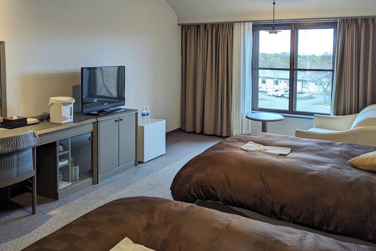 Une chambre d’hôtel comprenant un lit, un bureau avec une télévision, une chaise, une petite table et une fenêtre avec des rideaux.