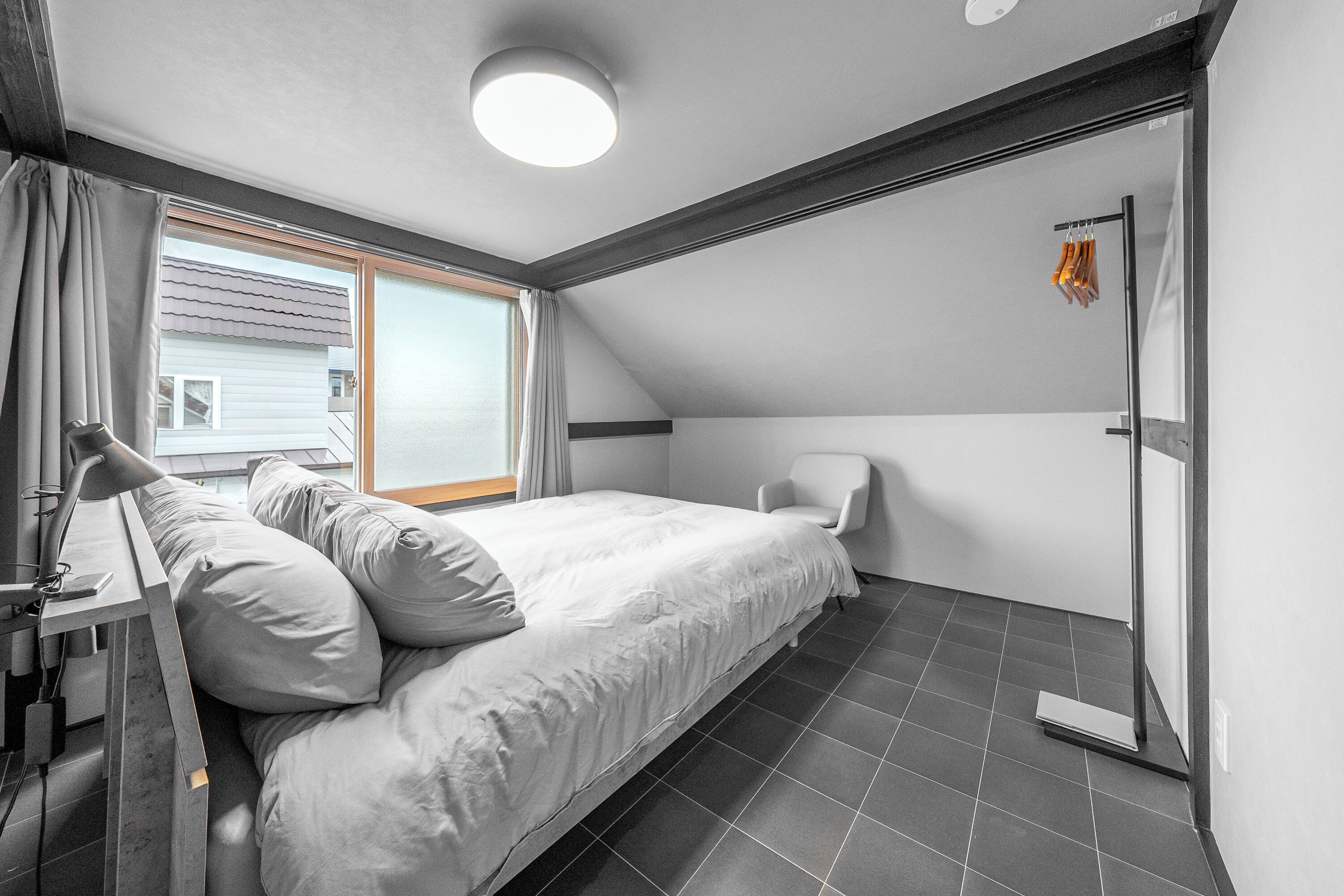 Ein Schlafzimmer mit Bett, Stuhl, Schreibtisch mit Lampe, Fenster mit Vorhängen und Tür mit Handtuchhalter.