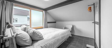 Ein Schlafzimmer mit Bett, Stuhl, Schreibtisch mit Lampe, Fenster mit Vorhängen und Tür mit Handtuchhalter.
