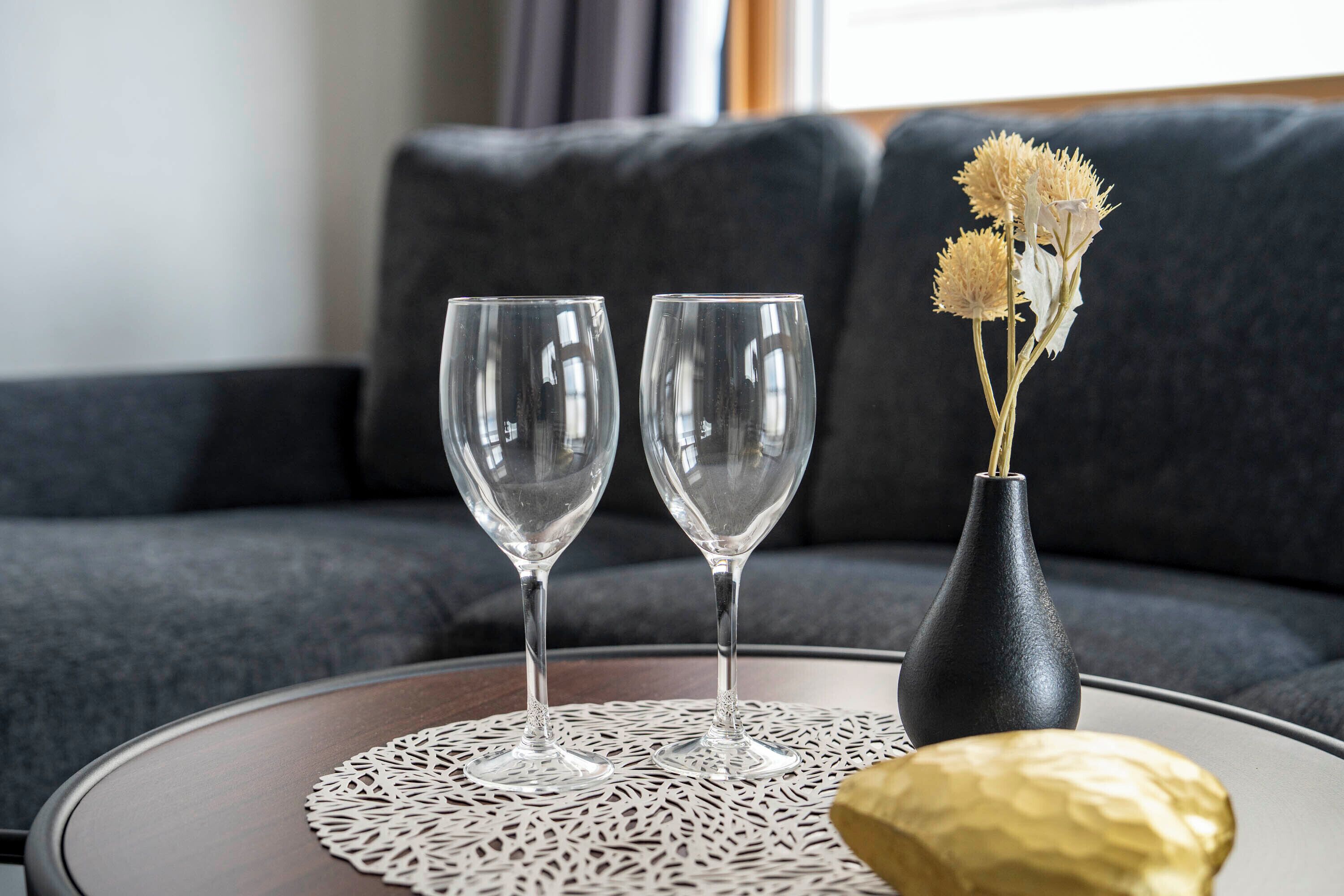 Two empty wine glasses on a round table with a doily, a vase with dried flowers, and a golden object, all set against a dark sofa in a living room.