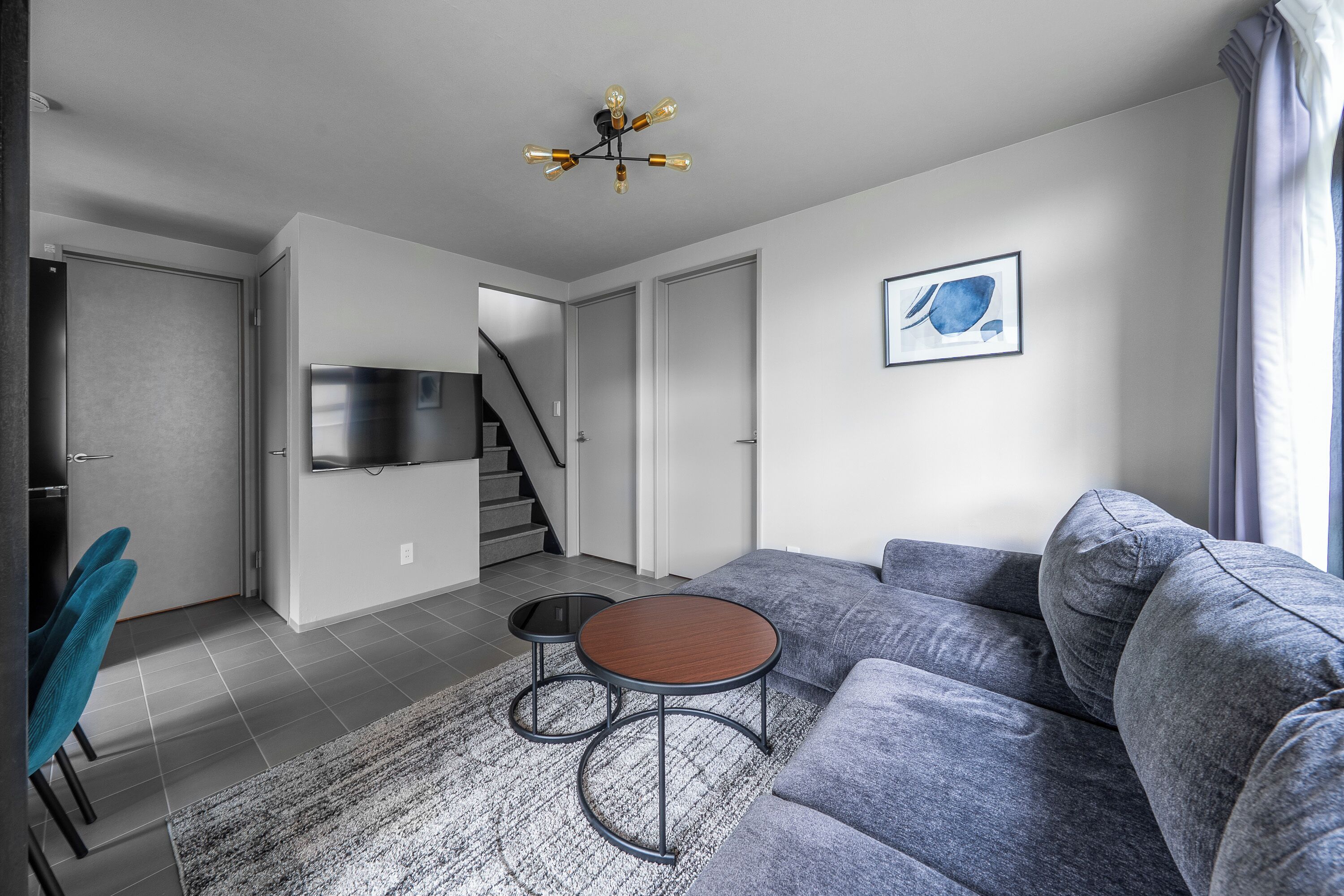Un saló modern amb un sofà gris, una taula rodona petita i una escala que condueix a una altra habitació.