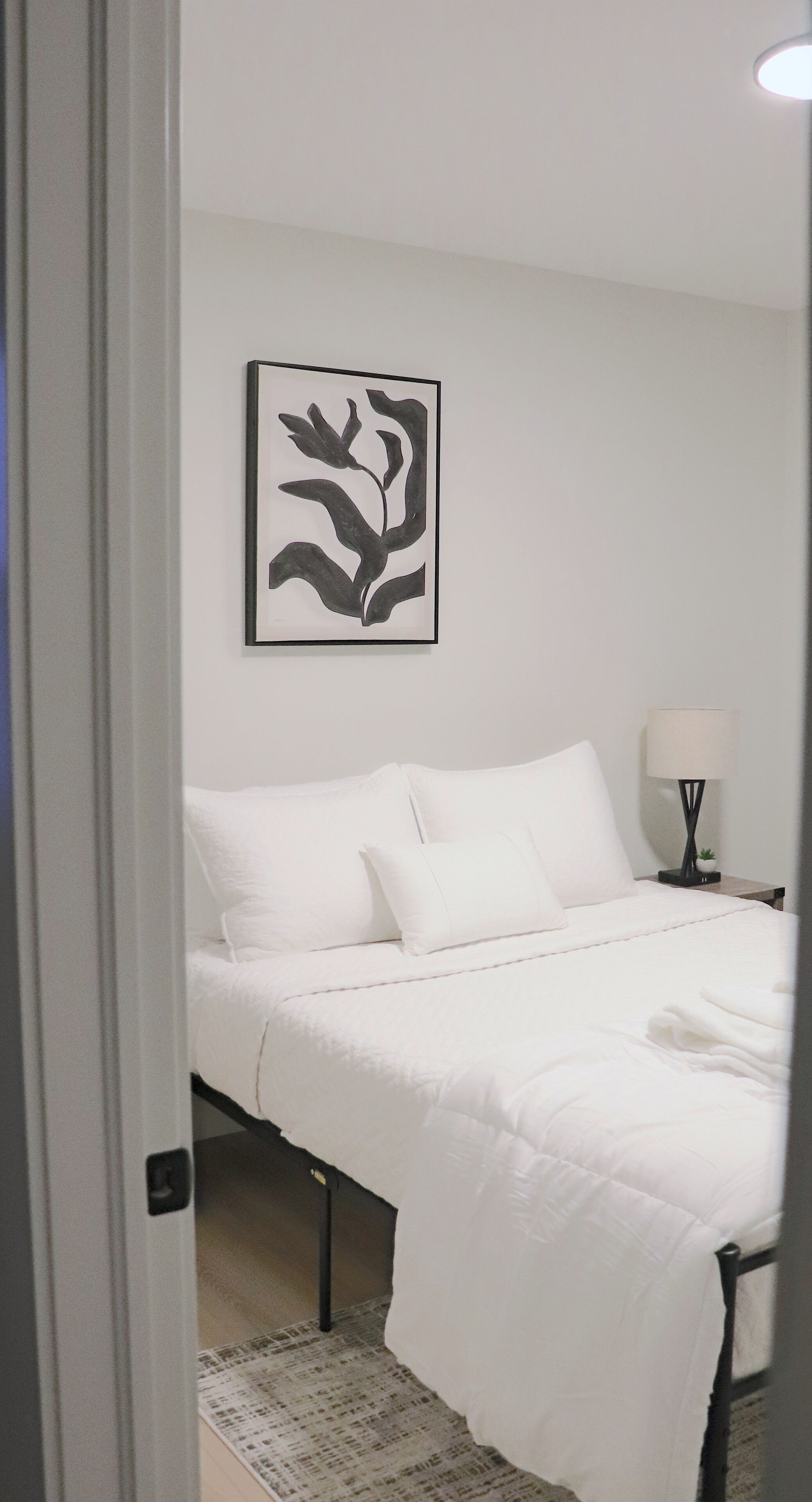 A neatly made bed with white bedding, a framed abstract art piece on the wall, and a bedside lamp.