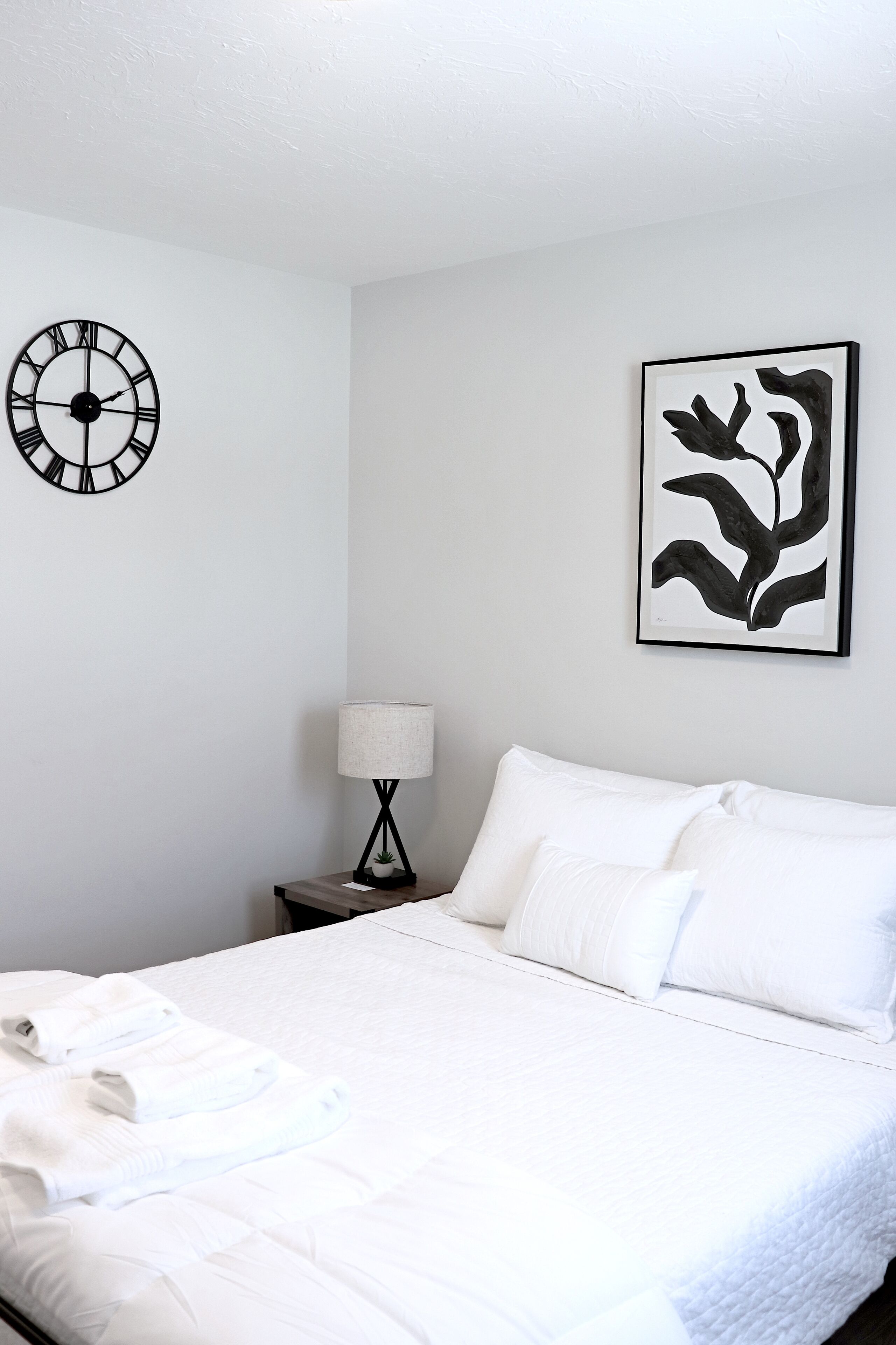 Un lit bien fait, avec du linge de lit blanc, une horloge murale, un tableau abstrait encadré et une lampe de chevet posée sur une table de nuit.
