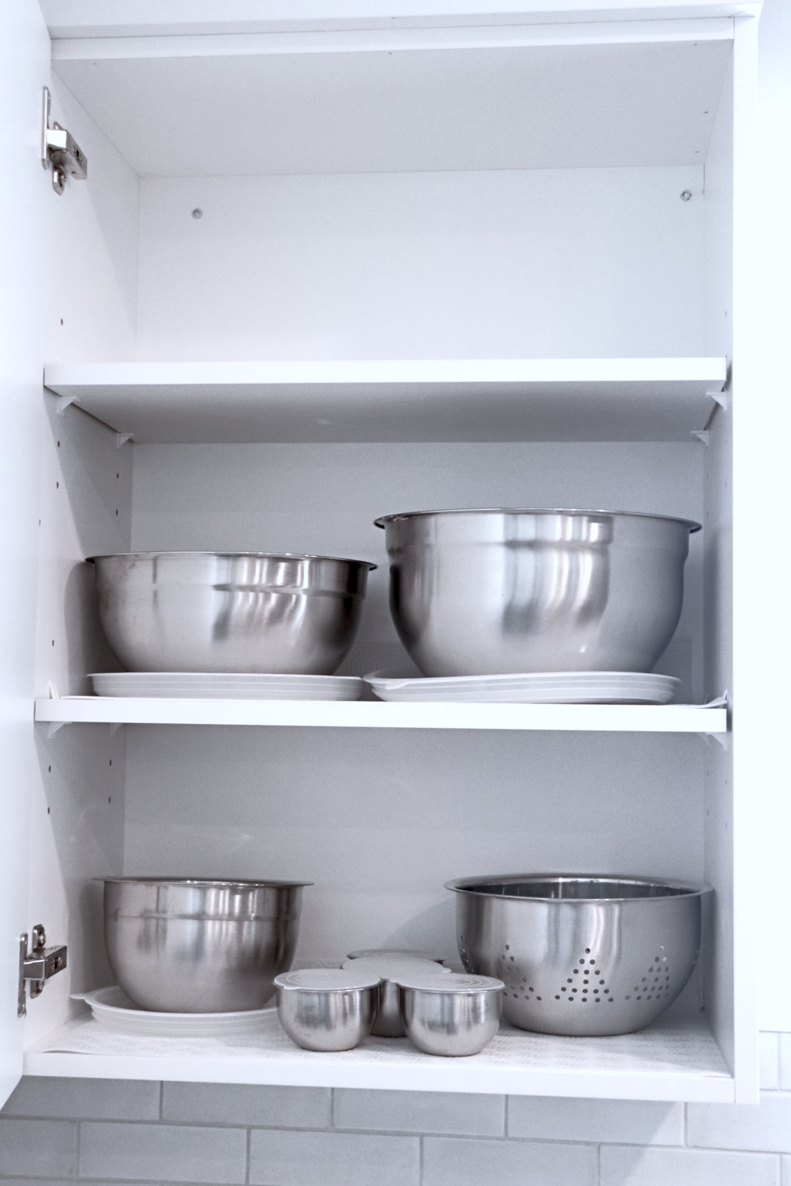 A kitchen cabinet with stainless steel cookware including a large pot, a smaller pot, a colander, and a mixing bowl.