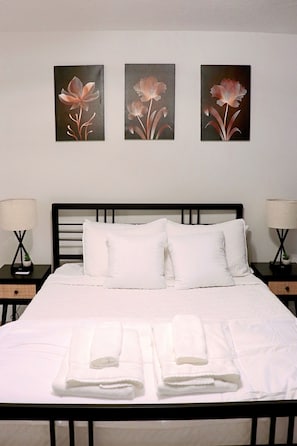 A hotel room with a bed, white bedding, two bedside tables with lamps, and three wall-mounted floral paintings. - New 2024 Home in Dieppe, NB (Dieppe)
