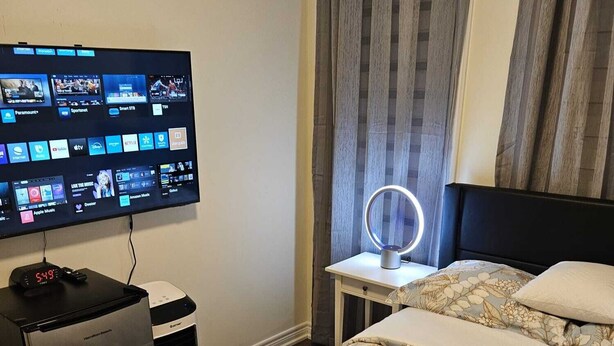 A hotel room with a bed, a desk with a lamp, a TV displaying various channels, and a heater. - Cozy clean, family sized house. (Cambridge)