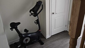 A room with a stationary exercise bike against a wall, a white door, and a wooden staircase. - Cozy clean, family sized house. (Cambridge)