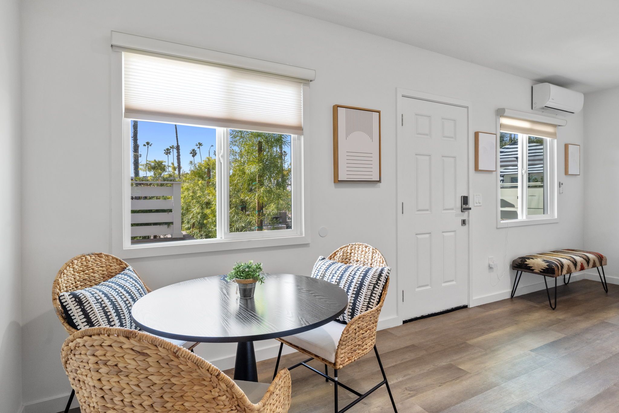 A room with a round table, wicker chairs, a potted plant, and a view of palm trees through the windows.