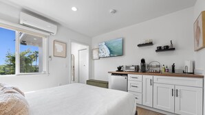 A bedroom with a bed, a small kitchenette, a window with a view of trees, and a mounted TV. - South Oceanside Bungalow 2 - Deluxe Queen Studio + Patio (Oceanside)