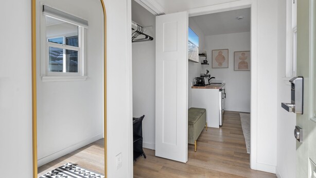 Een moderne, goed verlichte hal met een houten vloer, een witte deur en een uitzicht op een keuken met een bankje en kunstwerken.