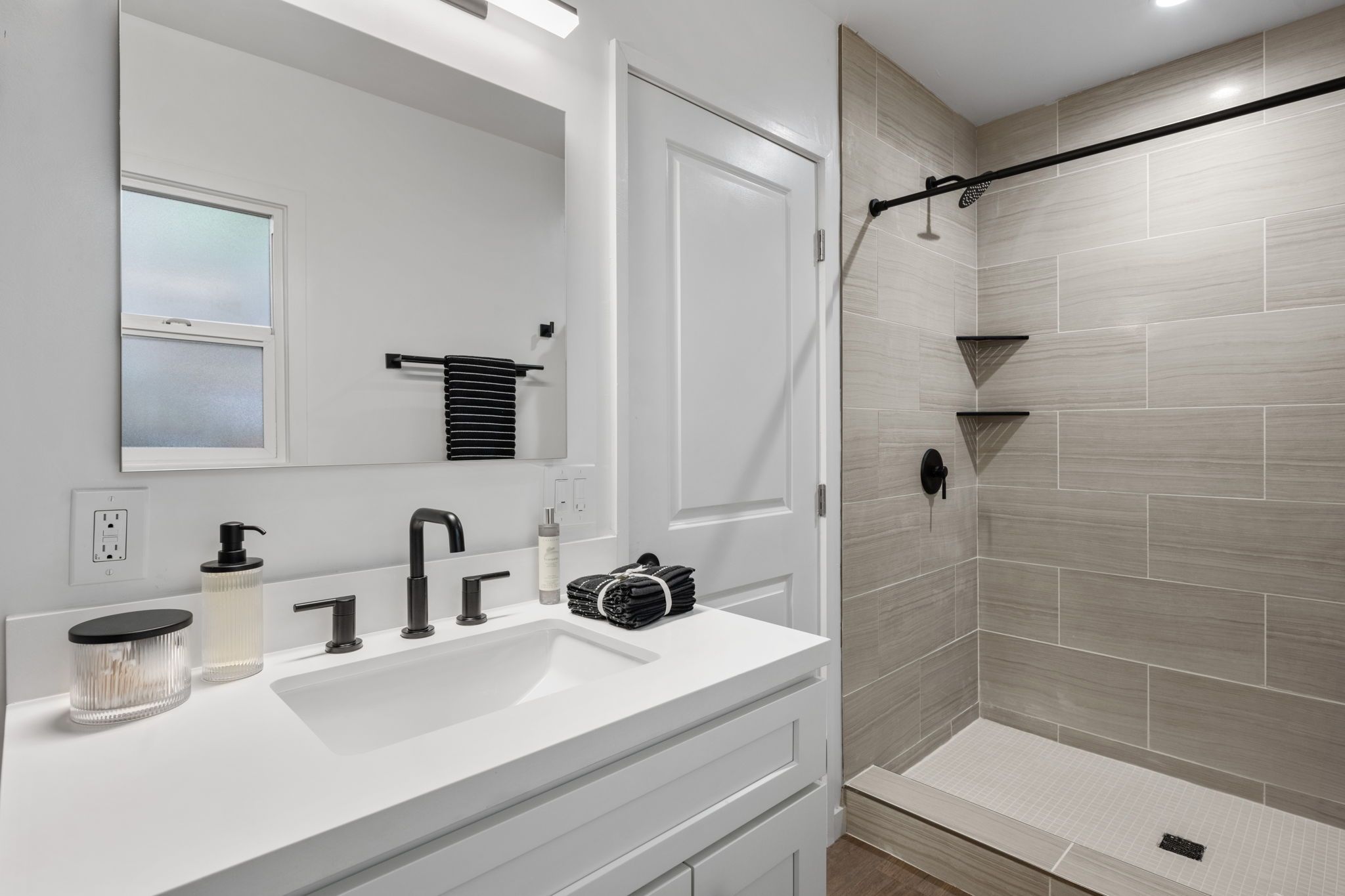 Un baño moderno con un tocador blanco, un espejo grande, un perchero y una ducha con grifo negro, además de paredes de ducha con azulejos.