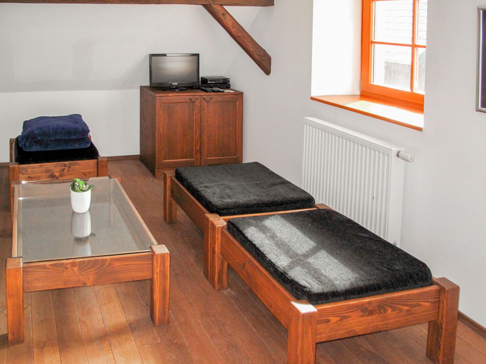 A small, cozy room with a wooden bench, a small table with a glass top, a television on a wooden cabinet, and a window with a view of the outside.