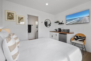 A modern bedroom with a large bed, a kitchenette, a dining area, and a beach view.