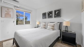 A bedroom with a large bed, two bedside tables with lamps, a window with a view, and wall-mounted artwork.