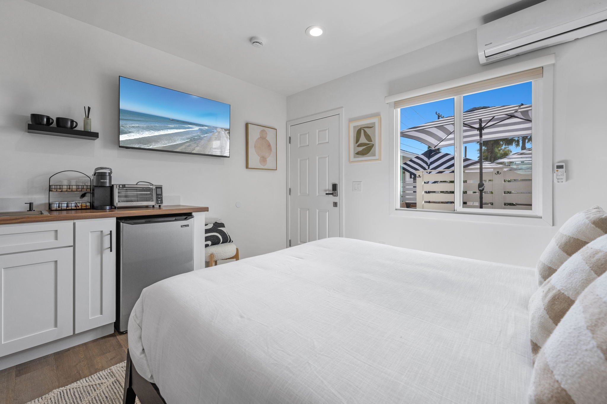 A bedroom with a bed, a kitchenette, a large window with a view of umbrellas and a beach, and a ceiling fan.