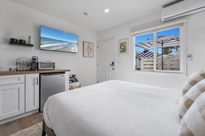 Een slaapkamer met een bed, een kitchenette, een groot raam met uitzicht op parasols en een strand, en een plafondventilator.
