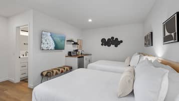 Quarto moderno com duas camas, uma kitchenette e uma TV exibindo uma cena de praia.