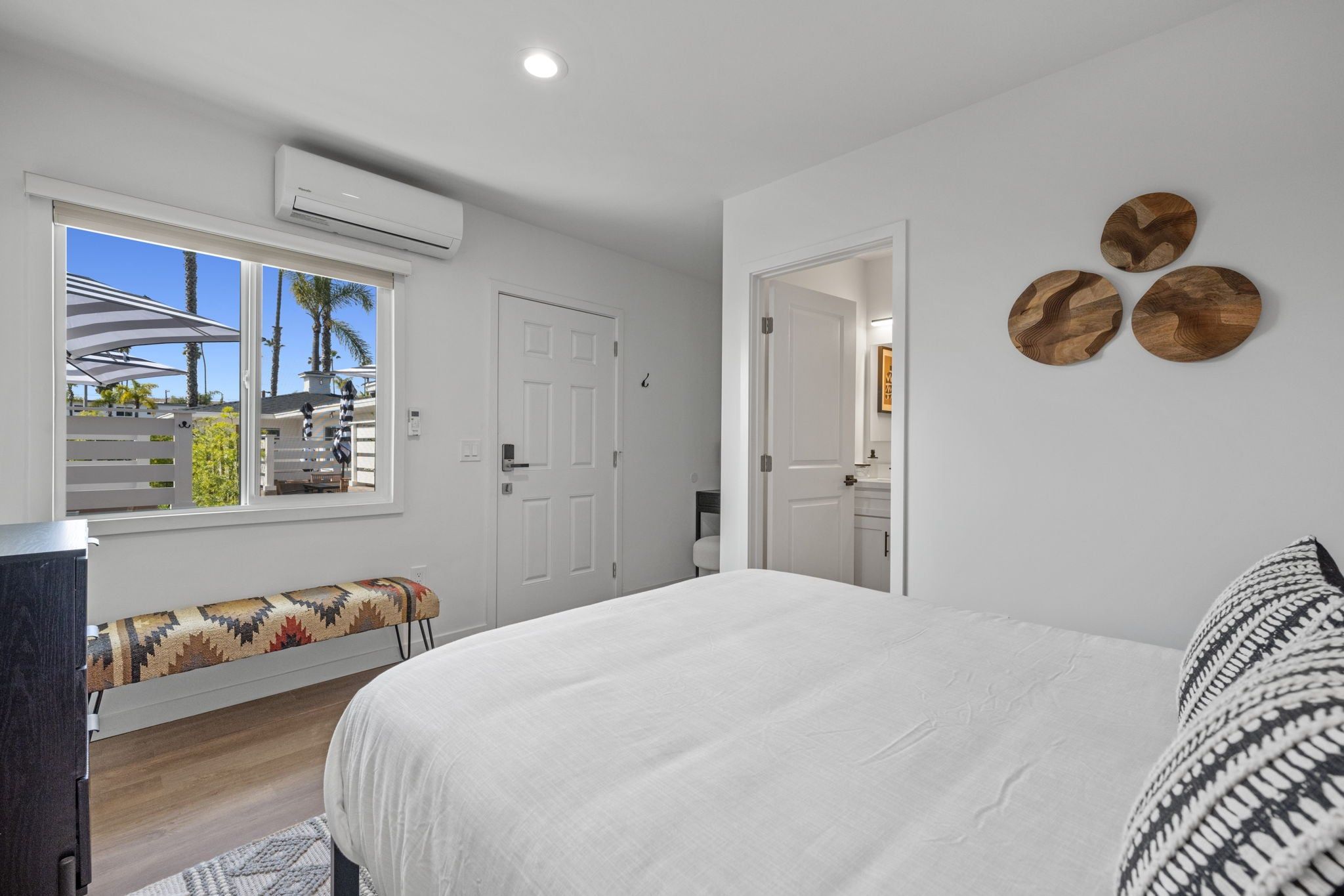 Ein Schlafzimmer mit einem Bett, einer Bank mit einem gemusterten Kissen, einem Fenster mit Blick auf Palmen und einer Wand-Klimaanlage.