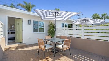 A patio with a table and chairs, two striped umbrellas, and a white fence.