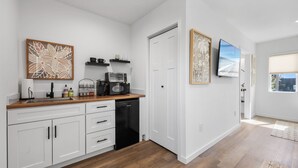 Eine moderne Küche mit weißen Schränken, einer Holz Arbeitsplatte, einem Geschirrspüler, einer Kaffeemaschine und einer Mikrowelle. An der Wand befinden sich zwei gerahmte Kunstwerke und ein Fernseher über der Arbeitsplatte.