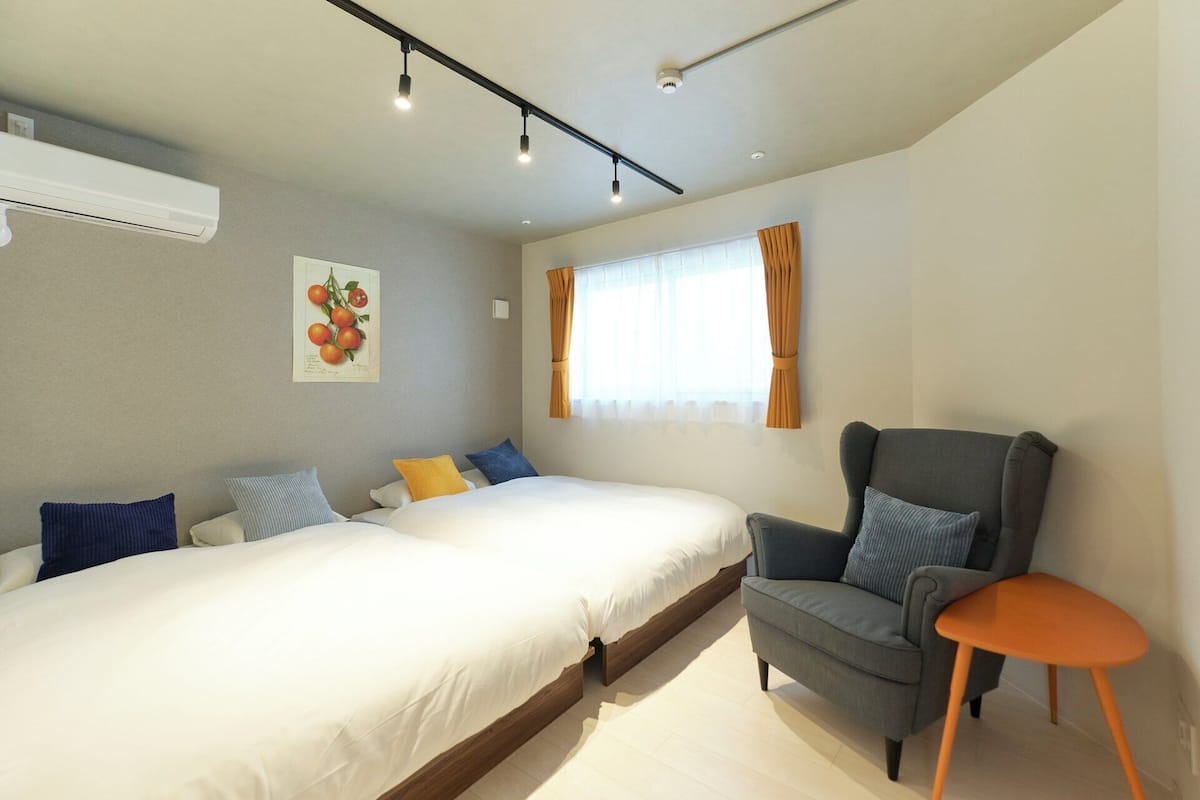 A modern bedroom with a large bed, a grey armchair, and a small orange table.