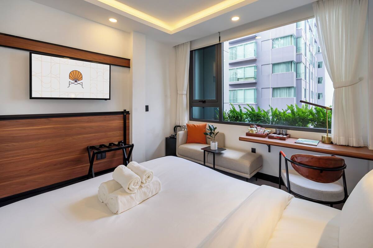 A hotel room with a large bed, a desk, a chair, a window with a view of a building, and a framed picture on the wall.