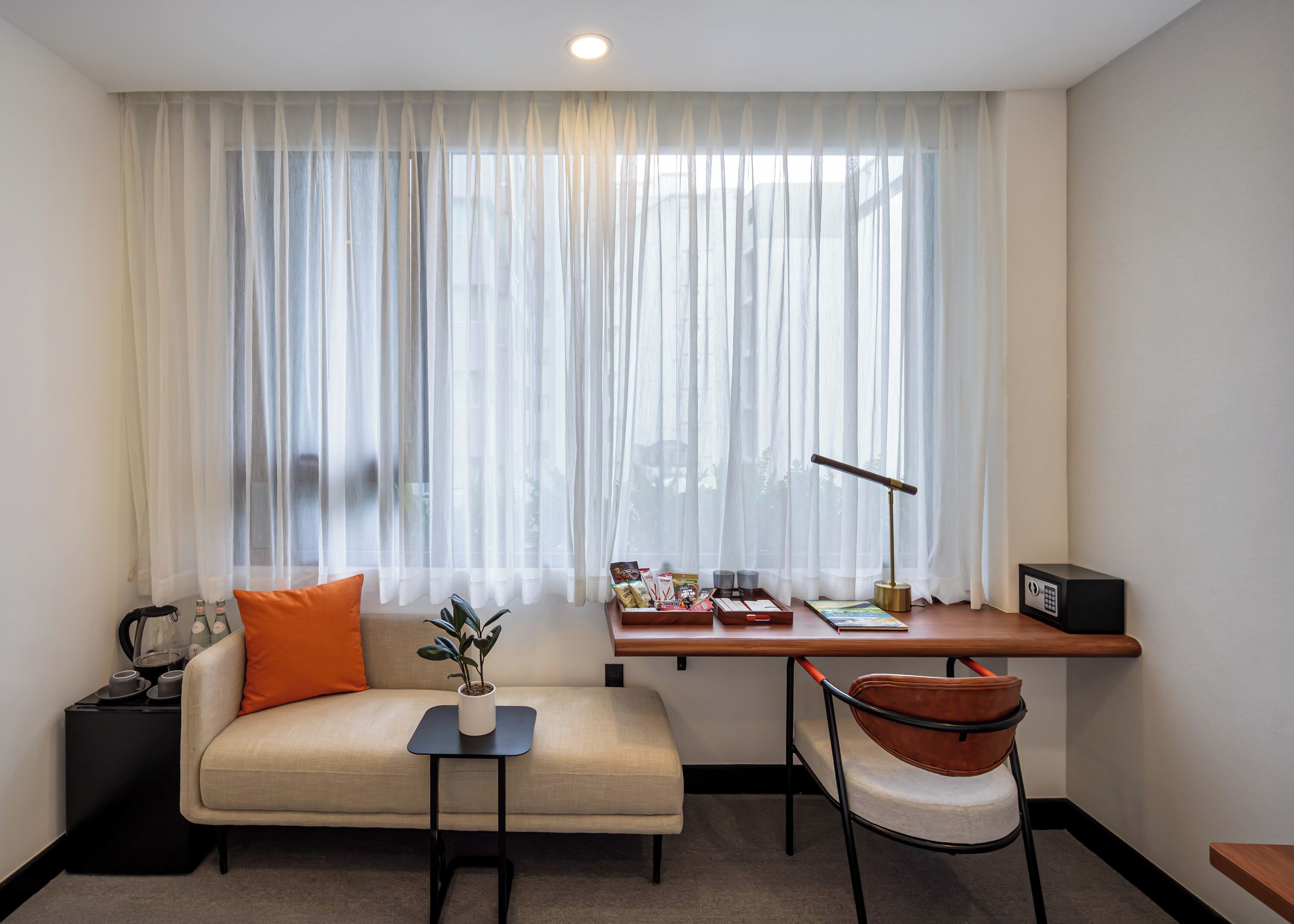 Ein modernes Hotelzimmer mit einer Couch, einem kleinen Tisch mit Pflanze, einem Schreibtisch mit Lampe und einem Sessel.