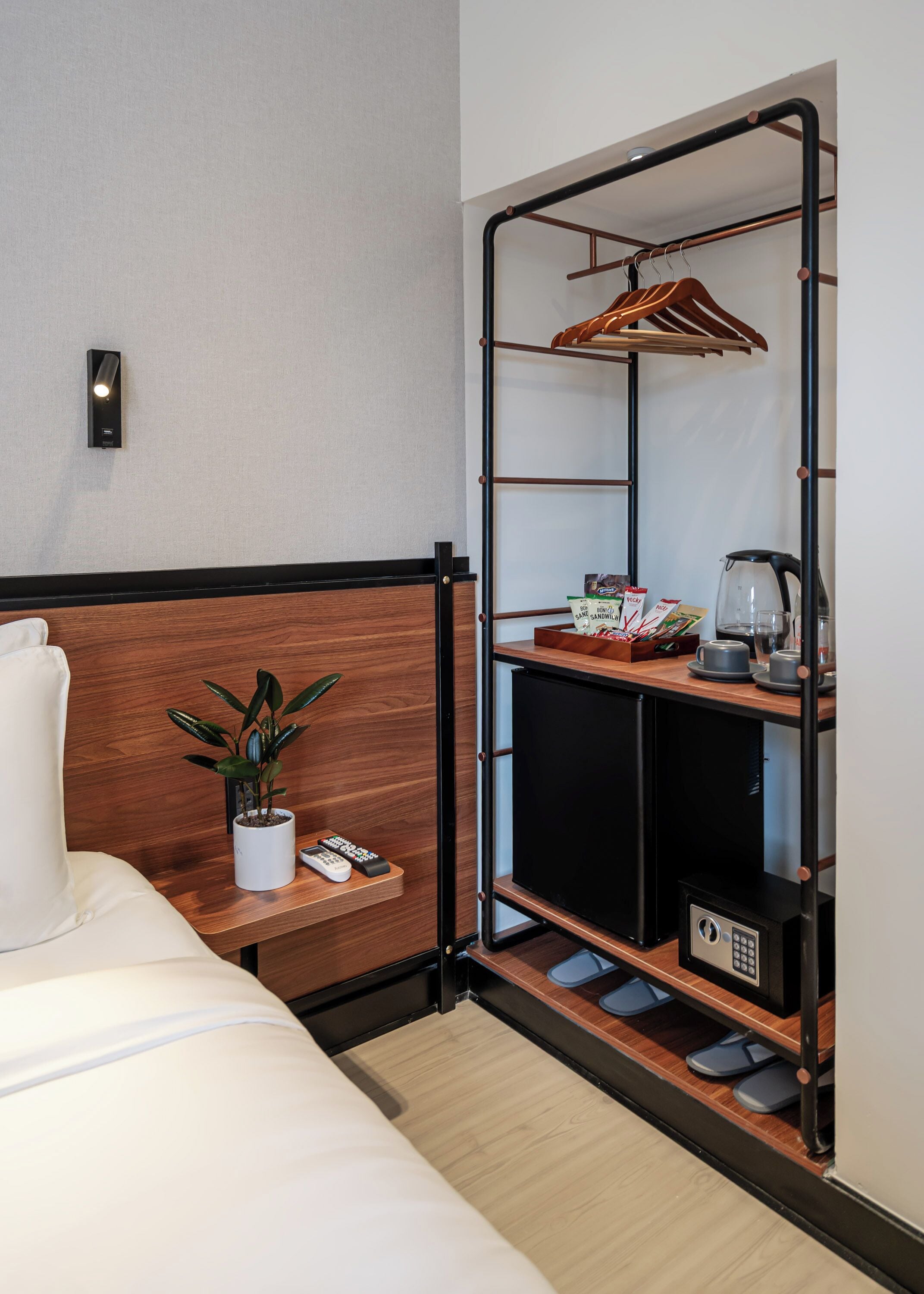 Habitación de hotel con cama, una mesita de noche con una planta, estante con hervidor, tazas y refrigerios, y un perchero con ropa.