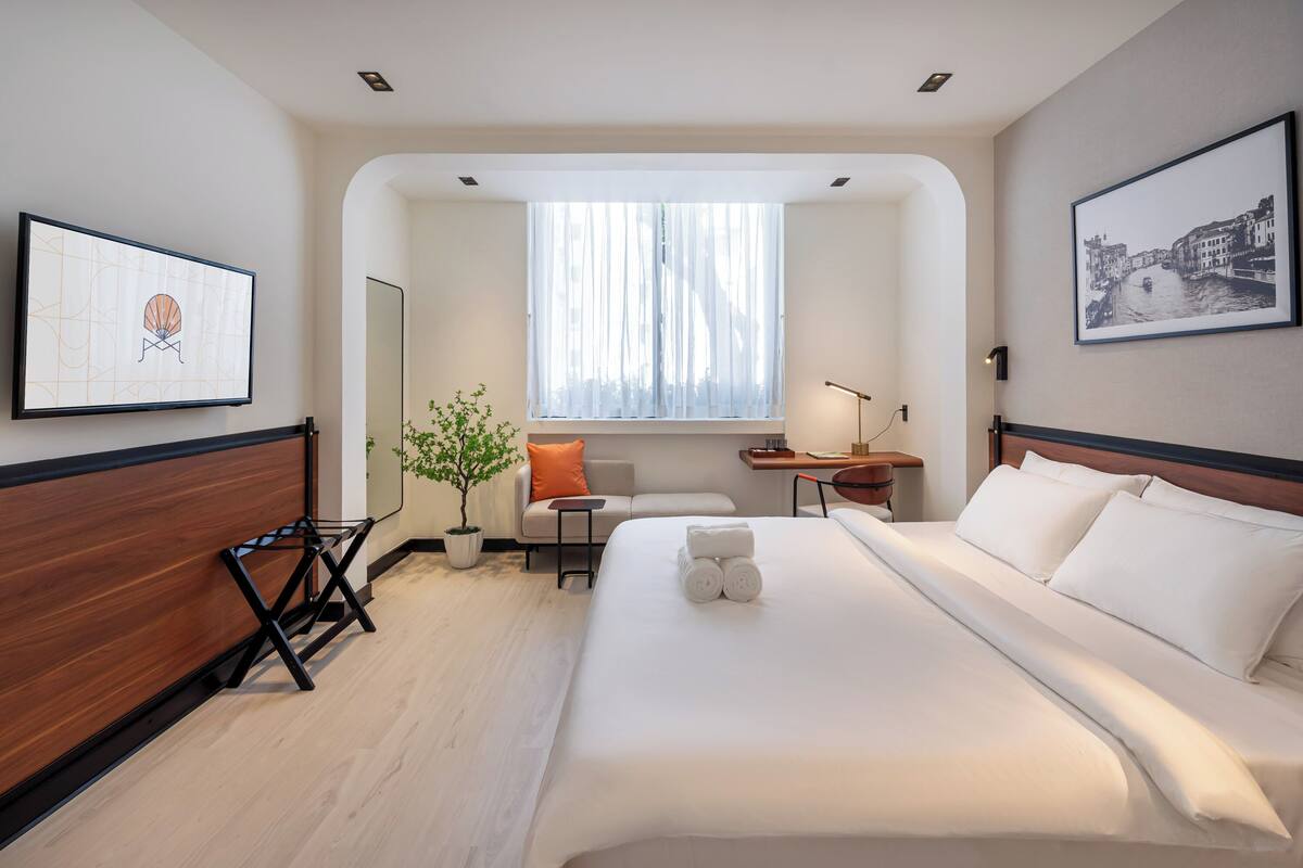 A modern hotel room with a large bed, a desk, a chair, a potted plant, and artwork on the walls.