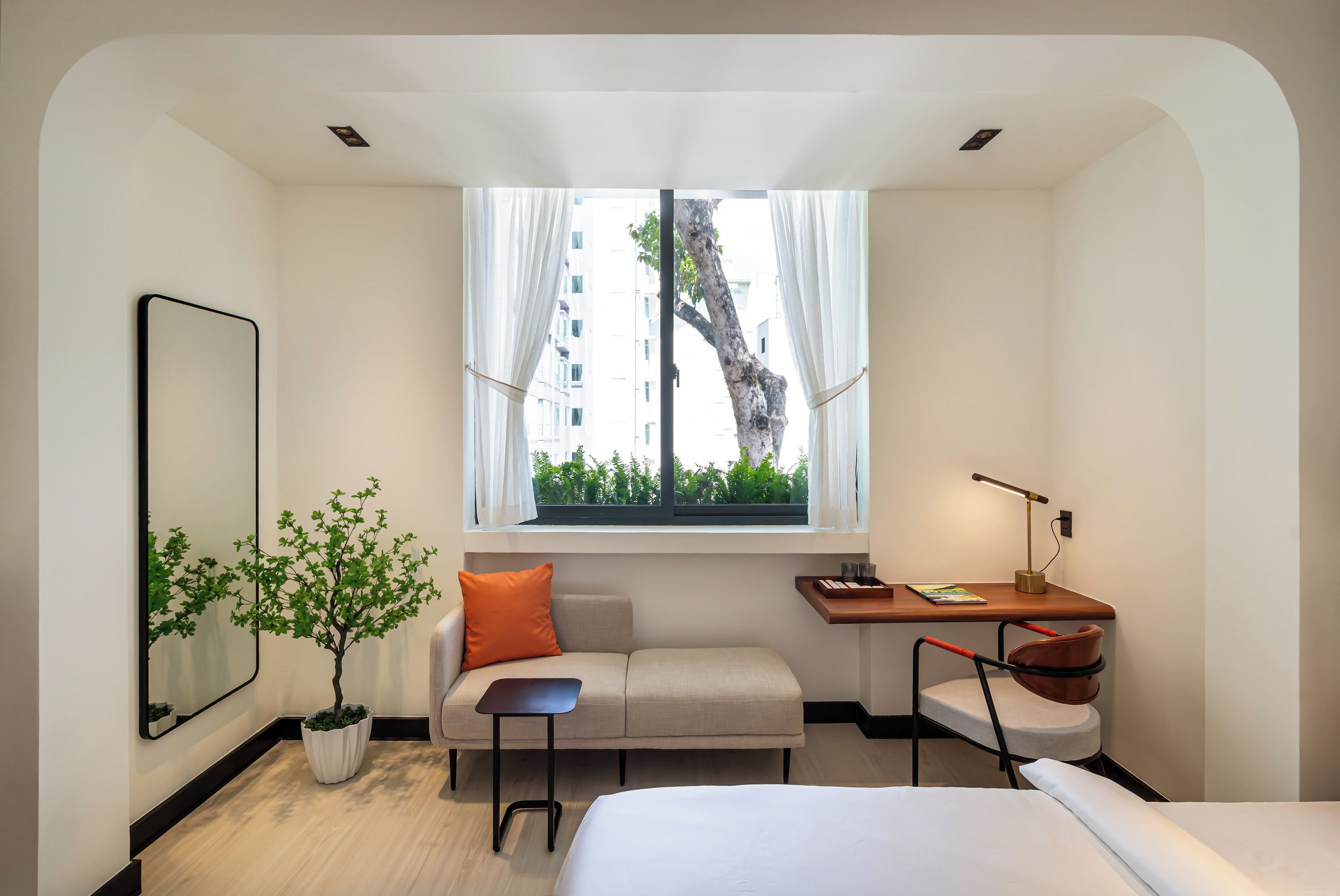 Una habitación de hotel moderna con cama, sofá, escritorio con silla, una mesita, planta en maceta y ventana con cortinas.