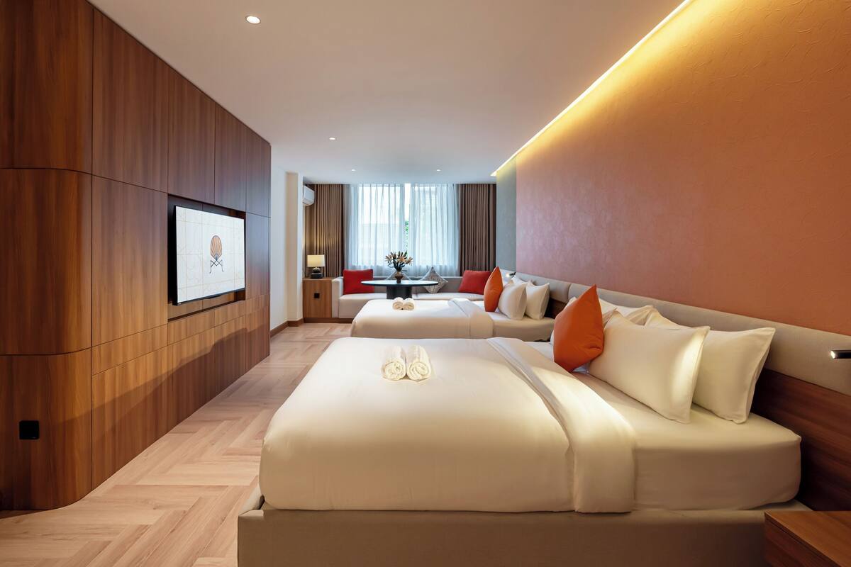 A modern hotel room with a wooden accent wall, a flat-screen TV mounted above the bed, a dining area with a table and chairs, and a large window with curtains.