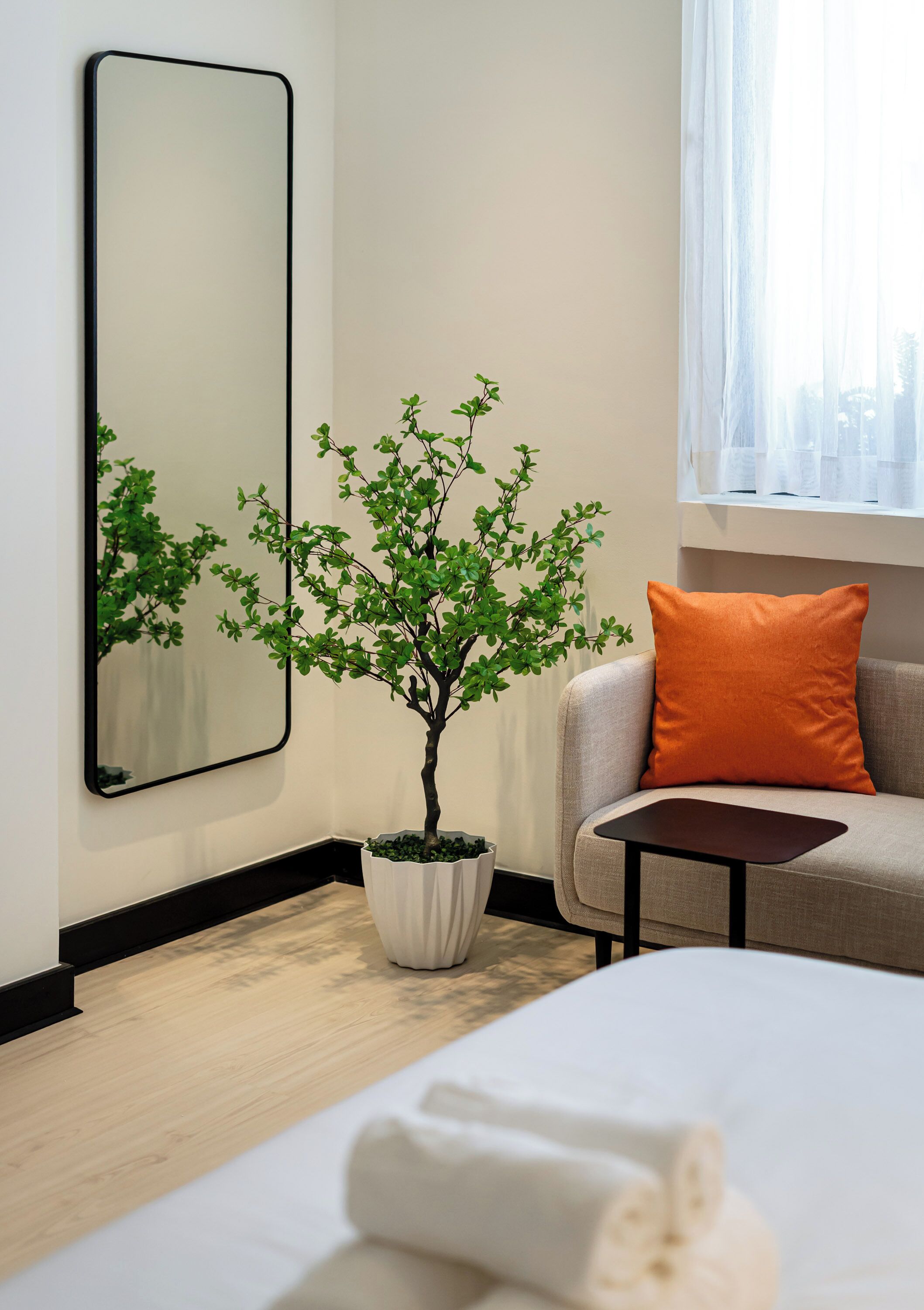 Habitación de hotel con cama, una mesita, un sofá con un cojín naranja, una planta en maceta y un espejo grande en la pared.