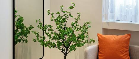 Habitación de hotel con cama, una mesita, un sofá con un cojín naranja, una planta en maceta y un espejo grande en la pared.