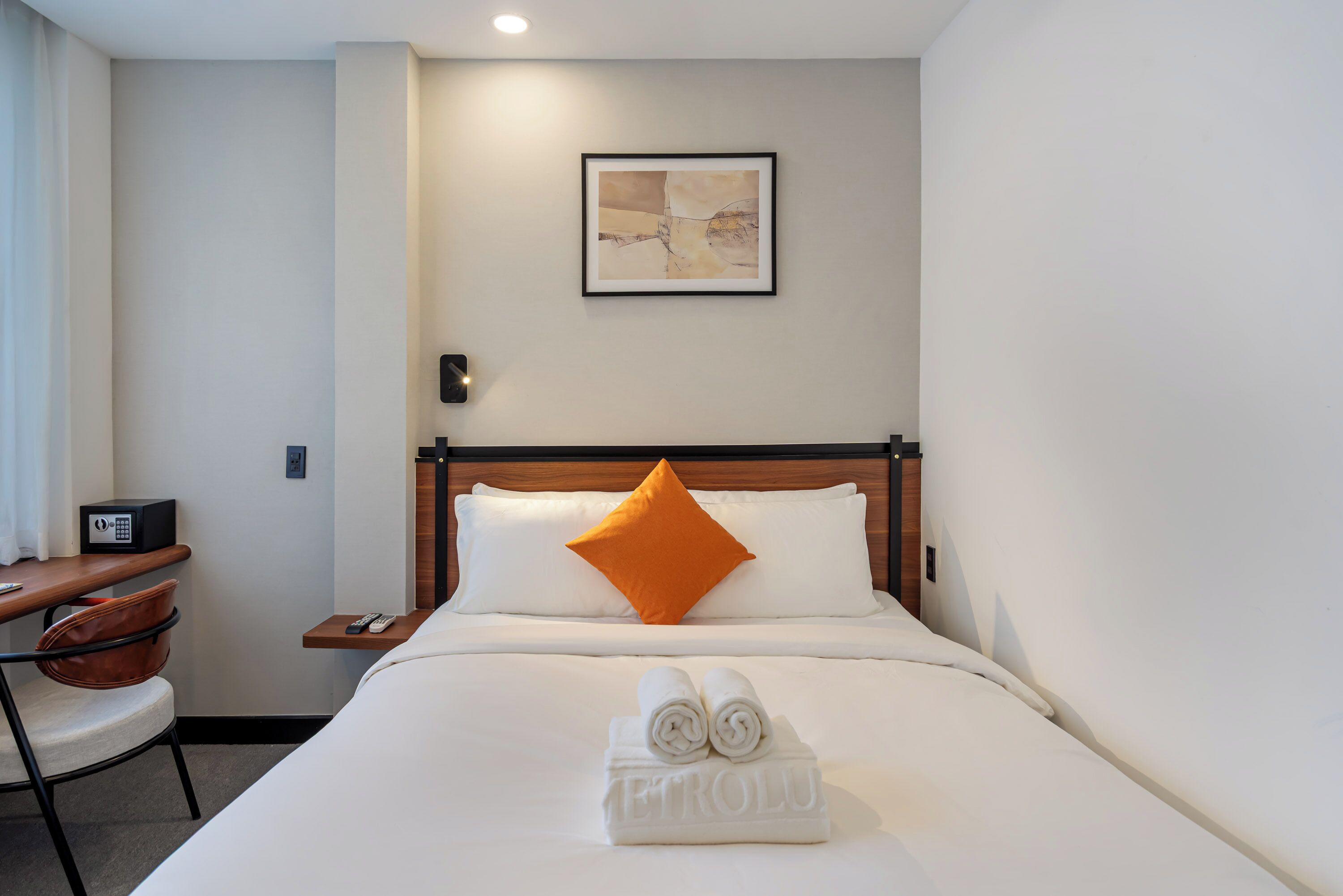 Quarto de hotel com uma cama, um travesseiro laranja, uma obra de arte emoldurada na parede, uma mesa com telefone e uma toalha dobrada na cama.