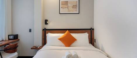 Quarto de hotel com uma cama, um travesseiro laranja, uma obra de arte emoldurada na parede, uma mesa com telefone e uma toalha dobrada na cama.