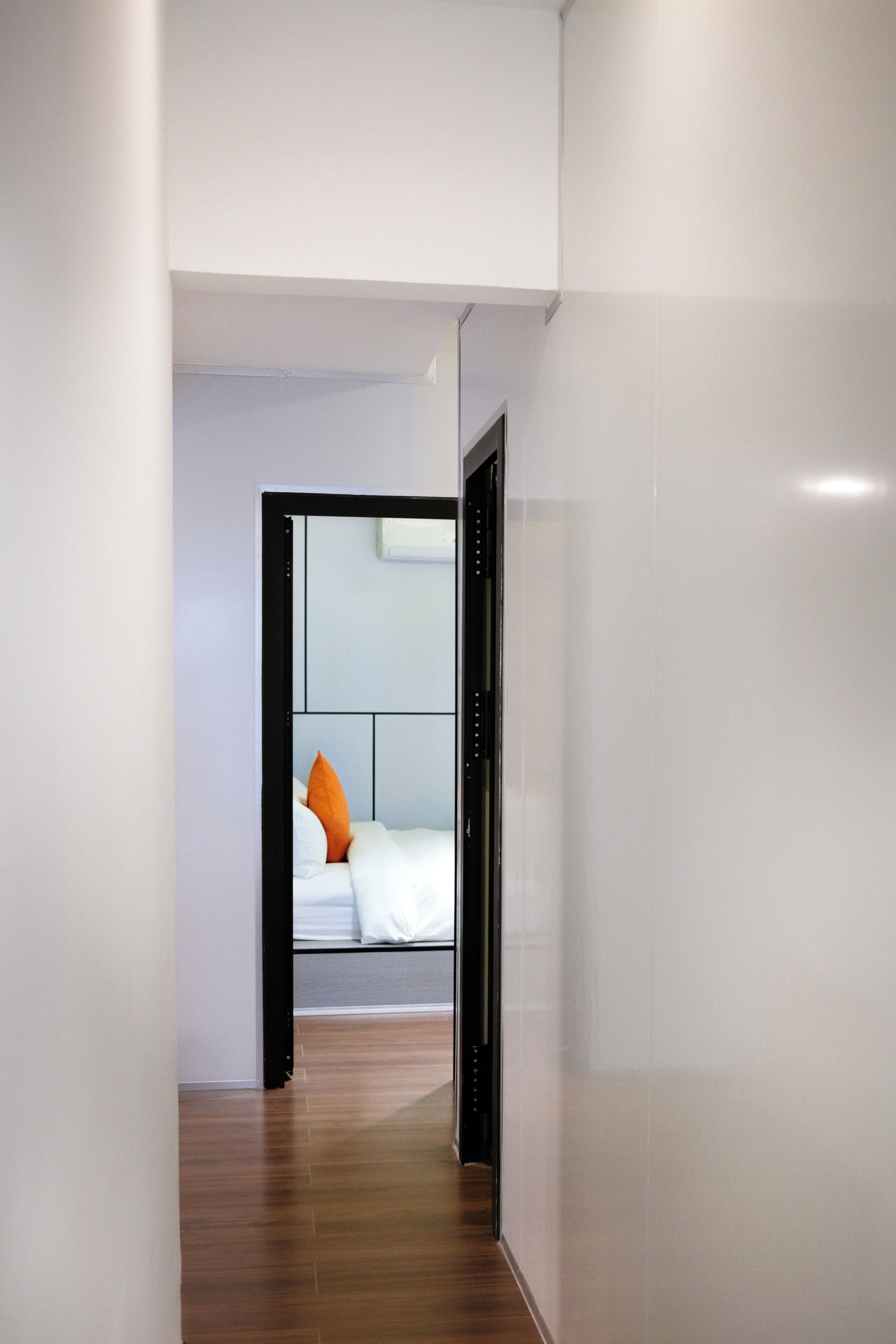 Un couloir d’hôtel moderne avec un plancher en bois, un lit avec une literie blanche et un oreiller orange, et une fenêtre avec des stores.