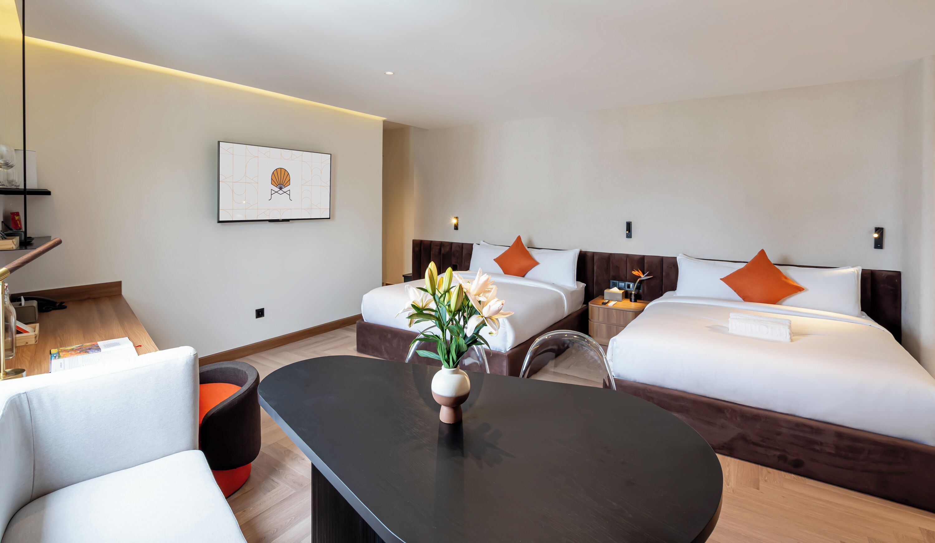 A hotel room with two beds, a desk, a chair, a vase with flowers, and a framed picture on the wall.