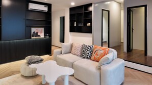 A modern living room with a sofa, a small white table, and a fluffy pouf. There is a black wall unit with shelves and a mounted air conditioner. - Metrolux City Contemporary Hotel (Ho Chi Minh City)