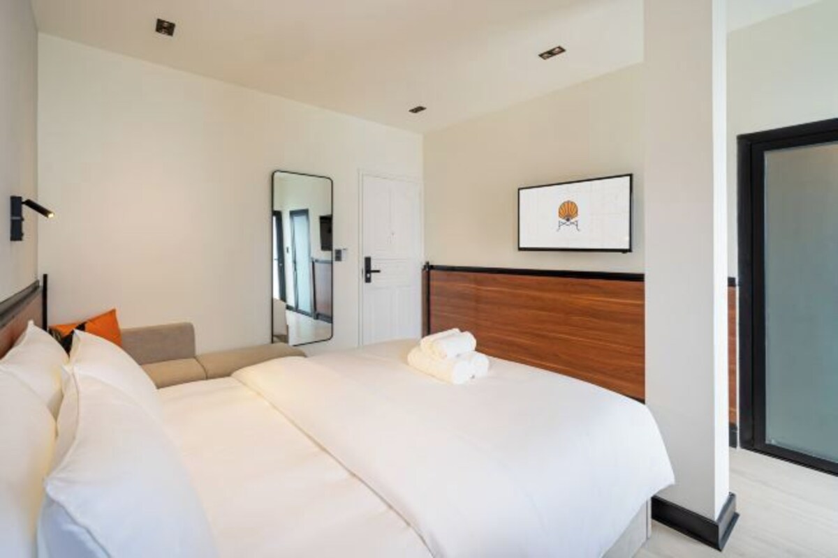 Quarto de hotel moderno com uma cama grande, cabeceira de madeira, um espelho, um quadro emoldurado e uma porta.