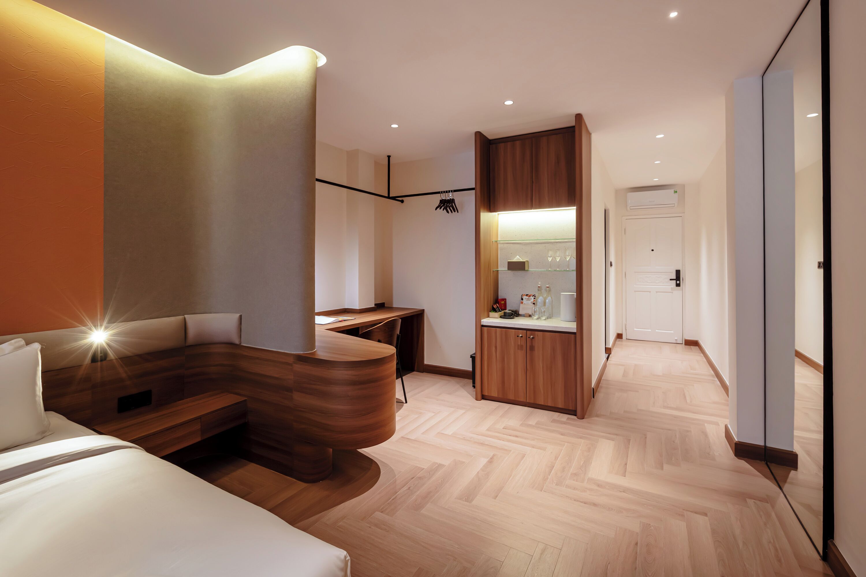 A modern hotel room with a curved bed, a wooden desk, a kitchenette, and a wardrobe.