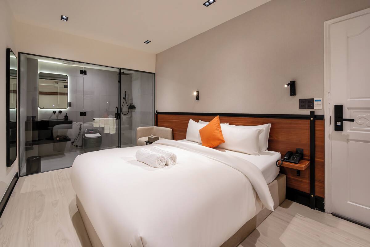 A modern hotel room with a large bed, a bedside table with a phone, a glass shower enclosure, and a wooden headboard.