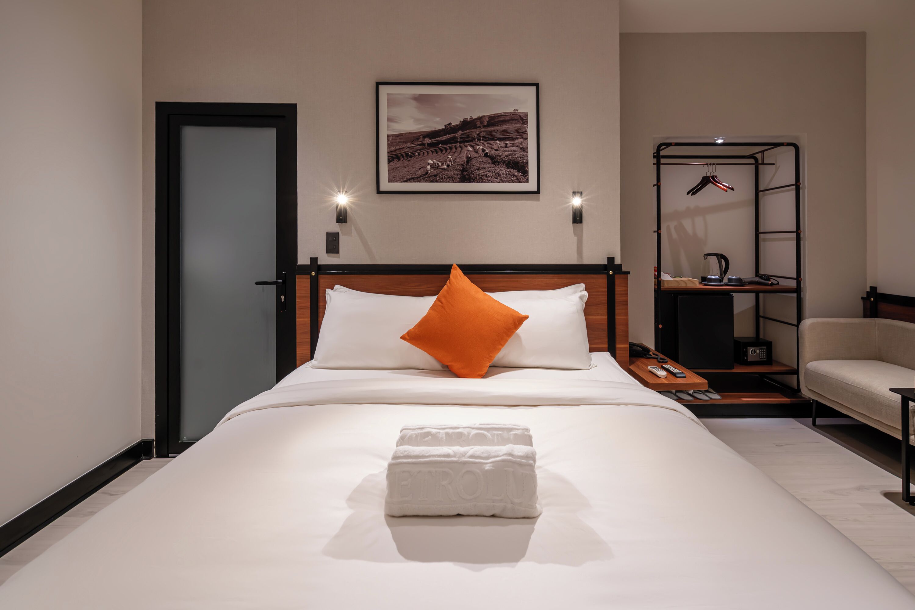 A neatly made bed with a white comforter and an orange pillow, a framed picture on the wall, a small shelf with a kettle, and a chair in a hotel room.