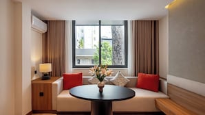 A modern hotel room with a round table, a sofa with red and patterned cushions, a vase of flowers, and a window with curtains. - Metrolux City Contemporary Hotel (Ho Chi Minh City)