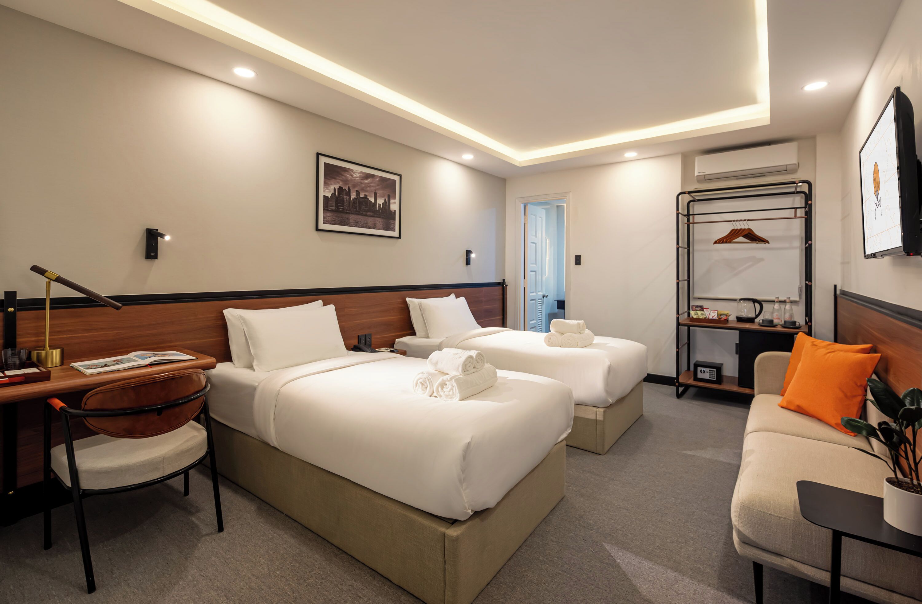 A hotel room with two beds, a desk, a chair, a sofa, and a wall-mounted TV.