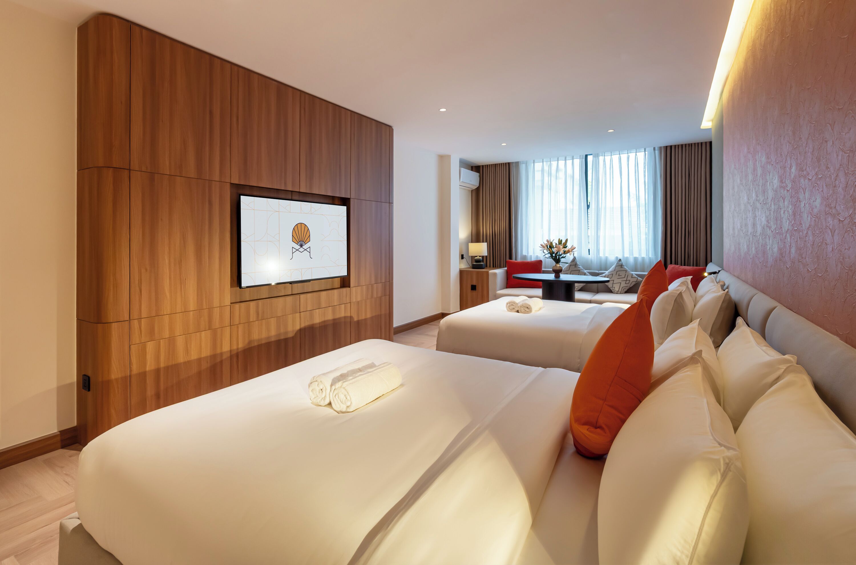 A hotel room with two beds, a wooden panel wall, a flat-screen TV, a dining area with a table and chairs, and a window with curtains.