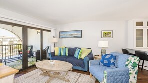 Uma sala de estar com um sofá azul, uma mesa de centro redonda de madeira e vista para o exterior por uma porta de vidro.