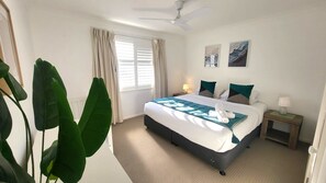 Una camera d'albergo con un letto, due comodini, una ventola a soffitto, una finestra con tende e una pianta.