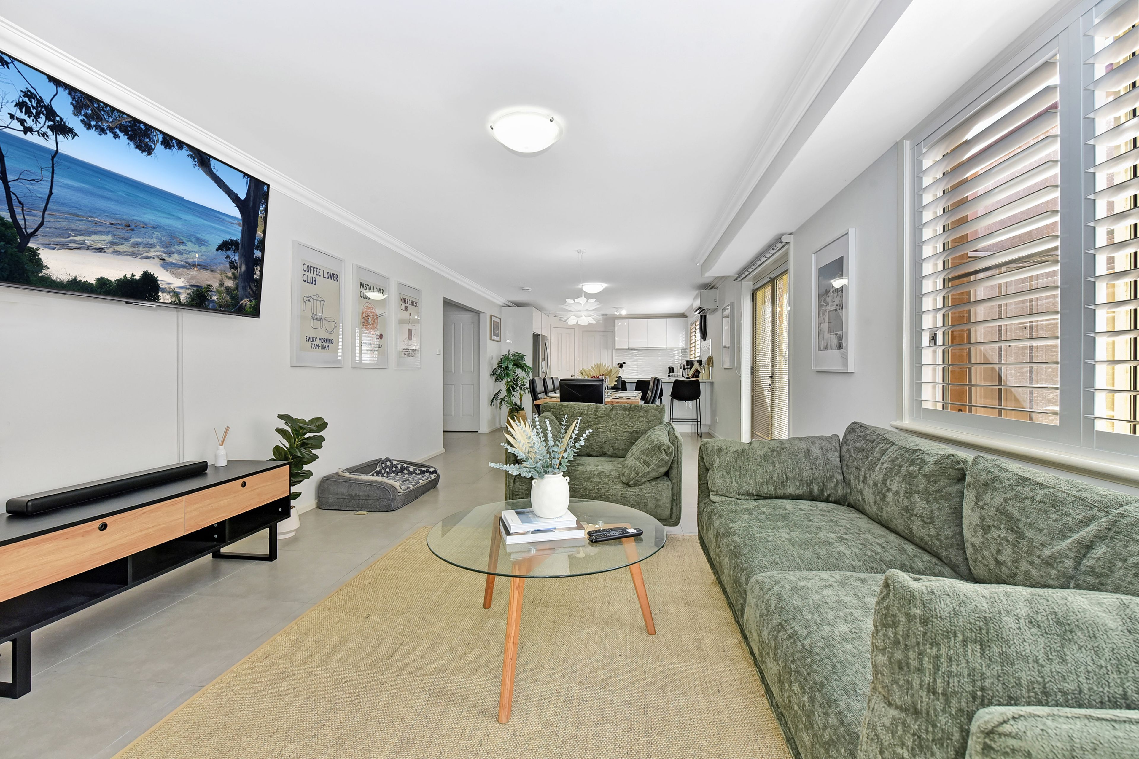 A modern living room with a green sofa, a glass coffee table, and a view of the kitchen.