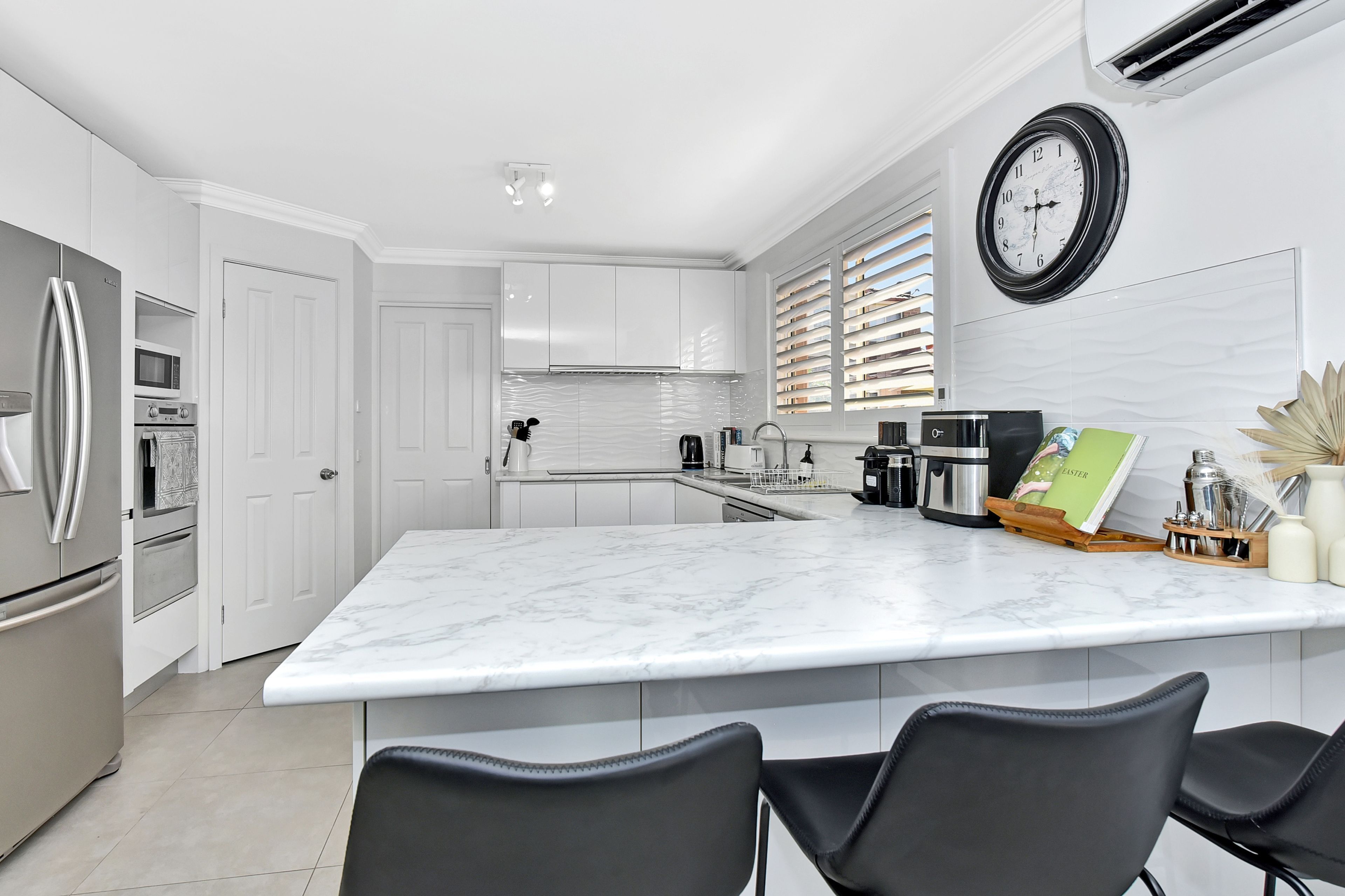Une cuisine moderne avec un îlot en marbre, des chaises noires et une horloge au mur.